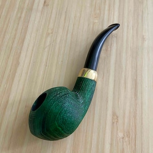 May include: A green wooden tobacco pipe with a curved black stem and a gold-colored band. The pipe has a classic shape with a bowl and a stem. The wood grain is visible on the surface of the pipe.