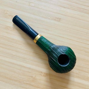 May include: A green and black wooden smoking pipe with a gold band. The pipe has a round bowl and a long, thin stem.