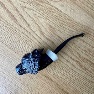 May include: A black tobacco pipe with a wolf head design. The pipe has a curved stem and a white band near the bowl. The wolf head is intricately carved with detailed features.