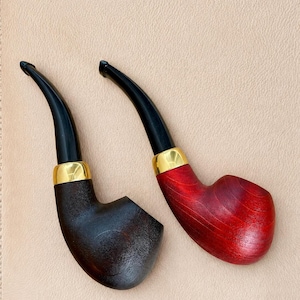 May include: Two tobacco pipes with curved black stems and gold-colored metal bands. One pipe features a dark brown, textured wooden bowl, while the other pipe has a smooth, vibrant red wooden bowl with a visible natural wood grain pattern.