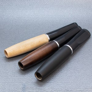 May include: Three wooden and black pens with silver accents. The pens are arranged in a triangular shape on a gray textured surface.