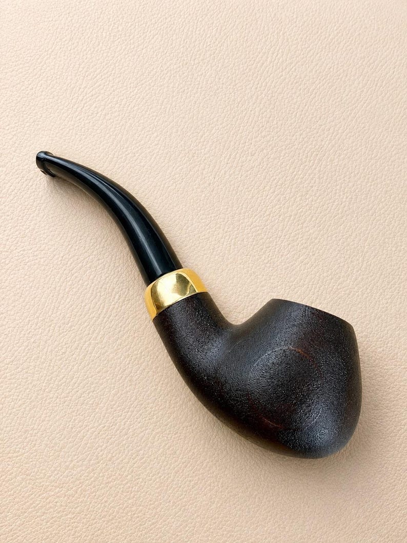 May include: A tobacco pipe featuring a dark brown, textured bowl and a smooth, curved black stem. The pipe is accented with a polished gold-colored metal band connecting the bowl to the stem, creating a distinct contrast between the materials.