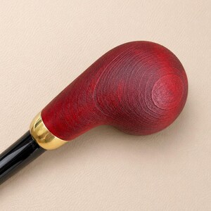 May include: A walking stick handle featuring a smooth, rounded, deep red stained wood top with visible grain patterns. A polished gold-tone metal collar connects the wooden handle to a sleek, glossy black shaft, creating a contrast in texture.