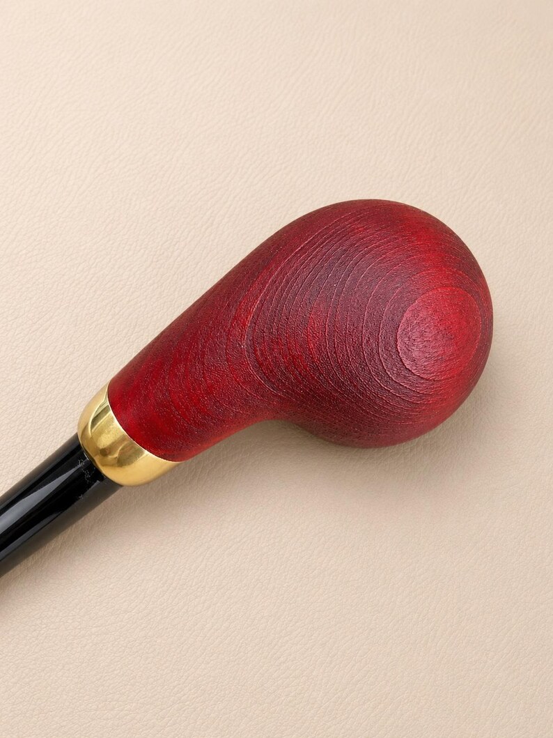 May include: A walking stick handle featuring a smooth, rounded, deep red stained wood top with visible grain patterns. A polished gold-tone metal collar connects the wooden handle to a sleek, glossy black shaft, creating a contrast in texture.