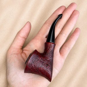 Puede incluir: Una pipa de tabaco con un hornillo de color rojo intenso y textura rústica, acompañada de una boquilla negra curva y lisa. La pipa presenta una forma angular distintiva con una base ancha que se estrecha hacia la boquilla, destacando el contraste entre la veta de madera oscura y rugosa y la boquilla de acrílico negro pulido.