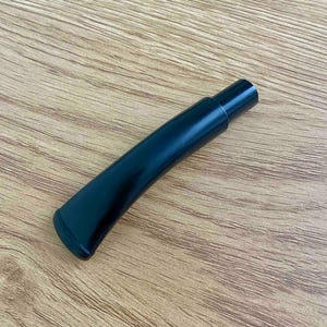 May include: A black, curved pipe stem with a glossy finish. The stem has a rounded end and a cylindrical mouthpiece. The pipe stem is set against a light brown wooden surface.