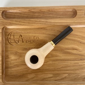 May include: A wooden pipe tray with a small, white pipe with a black bowl and a gold band. The tray has a logo that says "Anton & Co Pipes" with a globe design.