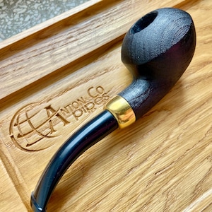 May include: A black wooden pipe with a gold band on the stem. The pipe is resting on a wooden tray with the text "Anton & Co. Pipes" engraved on it.