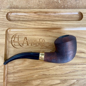 May include: A dark brown wooden pipe with a black mouthpiece and a gold band. The pipe is resting on a wooden tray with the text "Anton Co Pipes" engraved on it.