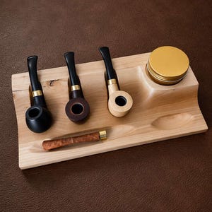 Puede incluir: Un soporte de madera clara con tres ranuras para pipas de tabaco con boquillas negras y anillos dorados, una pequeña herramienta marrón con patrón y un recipiente redondo con tapa dorada, todo sobre una base de madera rectangular.