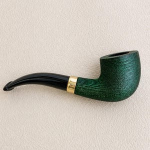 Puede incluir: Una pipa de tabaco con un hornillo de madera verde oscuro texturizado y veta visible. La pipa incluye una boquilla negra curva y lisa, conectada al hornillo mediante una banda de metal pulido de color dorado que crea un contraste definido.