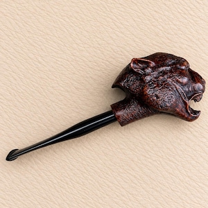 Puede incluir: Una pipa de tabaco con un hornillo texturizado de color marrón oscuro tallado con la forma de una cabeza de pantera rugiente con la boca abierta, unida a una boquilla negra lisa y cónica que se extiende desde la base de la figura felina.