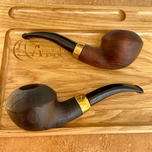 May include: Two wooden smoking pipes with black stems and gold accents on a wooden tray. The tray has the text "Anton Pipes" engraved on it.