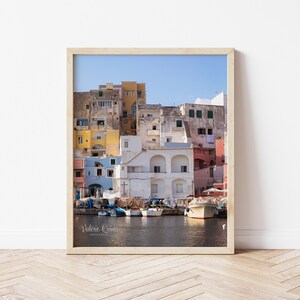May include: A colorful cityscape with buildings in shades of yellow, blue, pink, and white, overlooking a harbor with boats docked. The water is calm and reflects the sky, which is a clear blue.