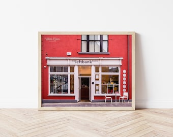 May include: A red brick building with a white storefront. The storefront has a sign that reads "the leftbank bistro". There are two tables and chairs outside the bistro.