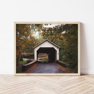 Puede incluir: Un puente cubierto blanco con un camino que lo atraviesa. El puente está rodeado de árboles con follaje otoñal. La foto fue tomada en un día nublado.