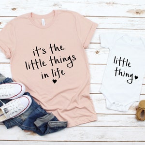 May include: A pink t-shirt with the text "it's the little things in life" and a small black heart. A white onesie with the text "little thing" and a small black heart. The shirts are on a white wooden background.
