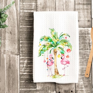 May include: A white kitchen towel with a pink flamingo and a palm tree decorated with Christmas lights and ornaments. The palm tree is decorated with yellow, red, and green ornaments.