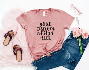 May include: A dusty rose t-shirt with the text "your custom design here" in black. The t-shirt is laid out on a white surface with a pair of pink shoes, a pair of glasses, a pair of jeans, and a bouquet of pink flowers.