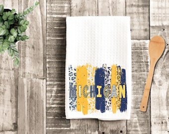 Op de afbeelding: Witte keukendoek met een abstract ontwerp in blauw en geel en het woord "MICHIGAN" in gouden letters.