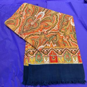 May include: A rectangular scarf featuring a colourful paisley design in red, orange, yellow, and black. The scarf has a navy blue border with a fringed edge. The scarf is displayed on a purple surface.