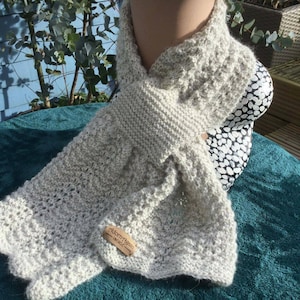 Puede incluir: Una bufanda de punto gris claro con un patrón delicado. La bufanda está drapeada sobre un maniquí y tiene una pequeña etiqueta marrón que dice "Aldermgrain Vintage".