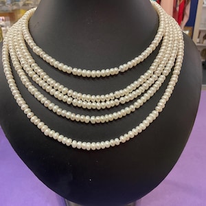 May include: A multi-strand pearl necklace displayed on a black mannequin. The necklace features several strands of small, round, white pearls, creating a layered effect. The pearls have a subtle luster, and the necklace is presented against a neutral background.