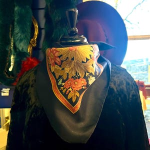 May include: A black silk scarf with a floral design in shades of yellow, orange, and brown. The scarf is displayed on a mannequin, with a dark hat in the background. The scarf has a red and orange border.