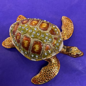 Pode incluir: Estatueta decorativa de tartaruga com corpo e membros dourados. A carapaça é um mosaico de verde, castanho e dourado, adornada com cristais transparentes. Em fundo roxo.