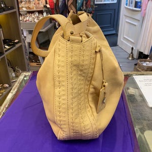 May include: A pale yellow leather shoulder bag featuring a textured surface with vertical rows of decorative whipstitch detailing. The bag includes a single shoulder strap, a hanging leather luggage tag, and a gathered top opening with metal hardware.