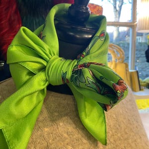 May include: A bright green silk scarf tied in a bow. The scarf features a floral pattern with red and green accents. The scarf is displayed on a black mannequin head, showcasing its shape and design.
