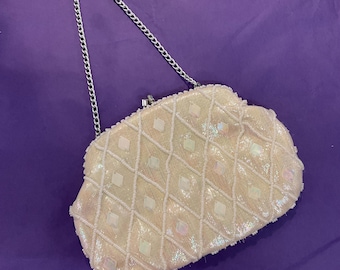 May include: A vintage cream-coloured beaded clutch purse with a silver chain strap. The purse features a diamond pattern with iridescent square accents. The clasp is silver, set against a purple background.