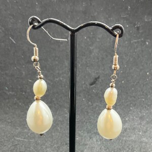 May include: Dangle earrings featuring a pearlescent white teardrop bead and a smaller oval bead. The earrings are suspended from silver-coloured fish hook ear wires. The earrings are displayed on a black earring stand.