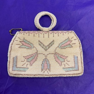 May include: Cream-coloured beaded handbag with a circular handle. The bag features a floral design in pink, blue, and silver beads. A small gold-coloured chain is attached to the side. The bag is set against a purple background.