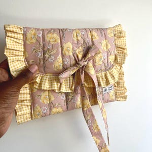 May include: A floral print clutch bag with a pink base and yellow flower pattern. It features a yellow and white gingham ruffle trim and a matching bow. The bag is held by a hand.