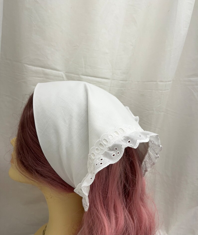 Puede incluir: Un pa&ntilde;uelo de cabeza de algod&oacute;n blanco con un borde de encaje blanco con ojales. El pa&ntilde;uelo est&aacute; atado alrededor de la cabeza de un maniqu&iacute;.
