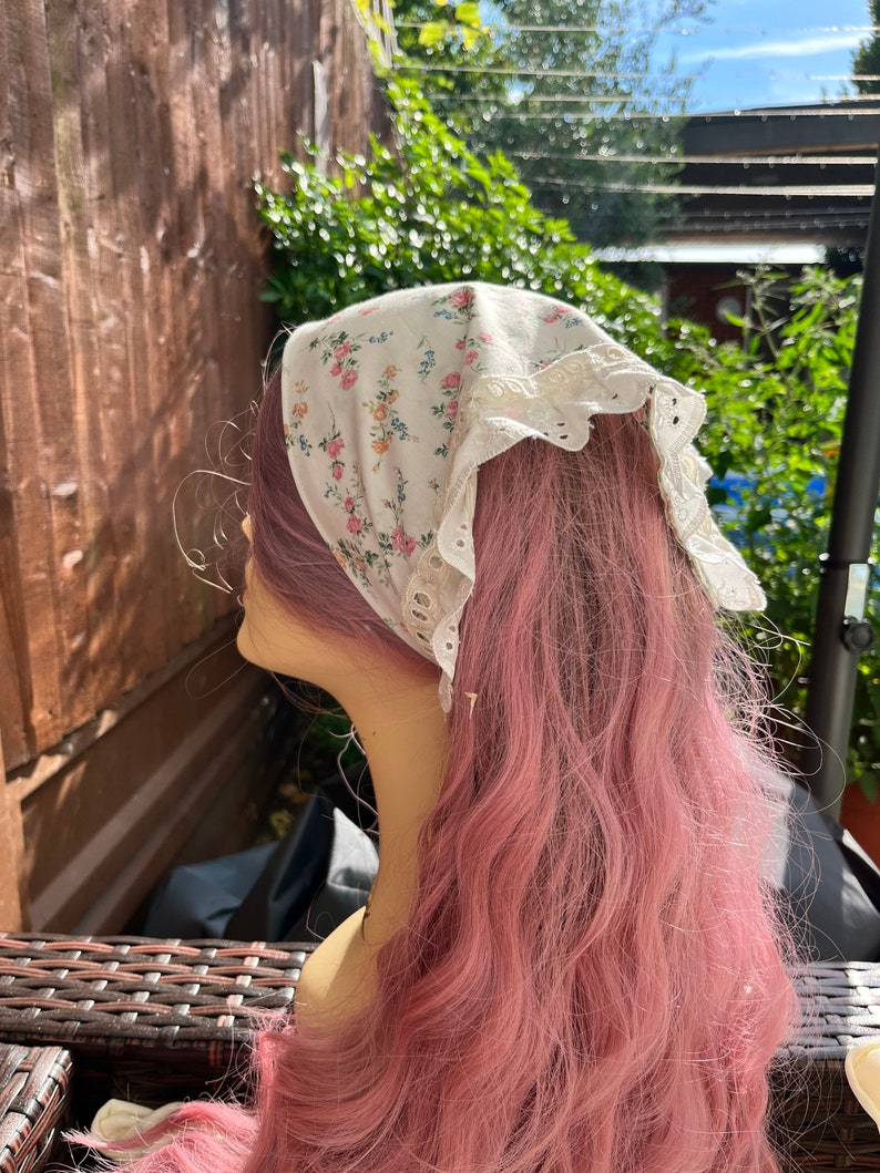 May include: A white floral patterned bandana with lace trim. The bandana is tied around a pink wig.