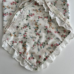 May include: A white cotton bandana with a floral print and white lace trim. The bandana is tied in a knot at the top.