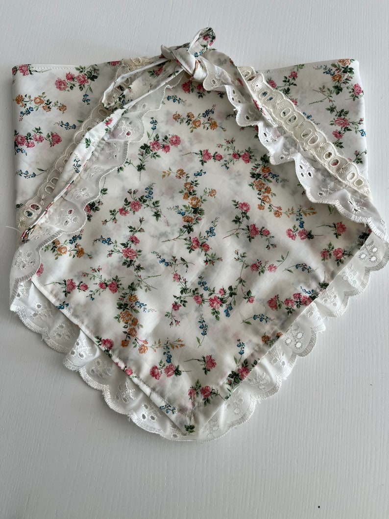 May include: A white cotton bandana with a floral print and white lace trim. The bandana is tied in a knot at the top.