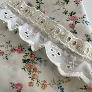 May include: White fabric with a floral print featuring small pink, blue, and yellow flowers. The fabric has a white lace trim with scalloped edges.