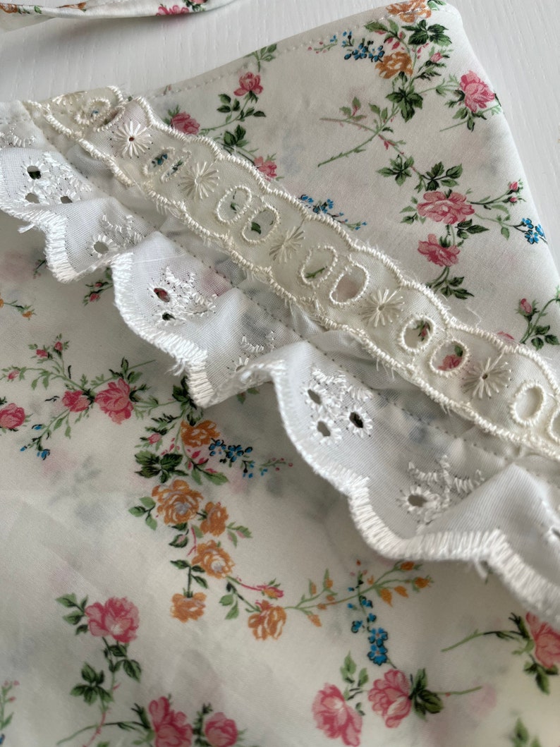 May include: White fabric with a floral print featuring small pink, blue, and yellow flowers. The fabric has a white lace trim with scalloped edges.