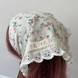 May include: A white cotton headscarf with a floral print and white lace trim. The scarf is tied on a pink wig.