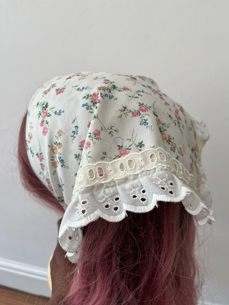 May include: A white cotton headscarf with a floral print and white lace trim. The scarf is tied on a pink wig.