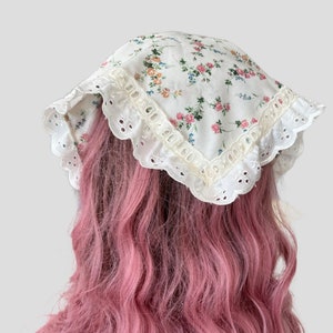 May include: A white cotton headscarf with a floral print and white lace trim. The scarf is tied on a person's head with pink hair.