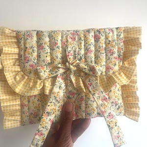 May include: A floral print clutch bag with a yellow gingham ruffle trim and a matching bow closure. The quilted fabric features a pattern of small pink and yellow flowers on a cream background. The bag is held by a hand.