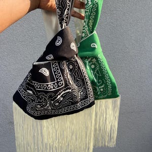 May include: Two knot bags made from black and green bandana fabric featuring a white paisley print. Each bag has a single handle formed by the fabric knot and is finished with long, cream-coloured fringe hanging from the bottom edge of the pouch.