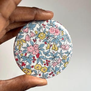 May include: A round, white fabric mirror with a floral pattern in shades of pink, yellow, blue, and red. The fabric has a vintage feel and is covered in a variety of small flowers and leaves.