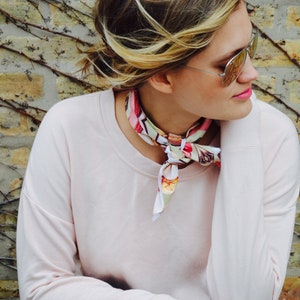 May include: A woman wearing a pink sweatshirt with a floral patterned scarf tied around her neck. She is wearing blue jeans and has blonde hair.