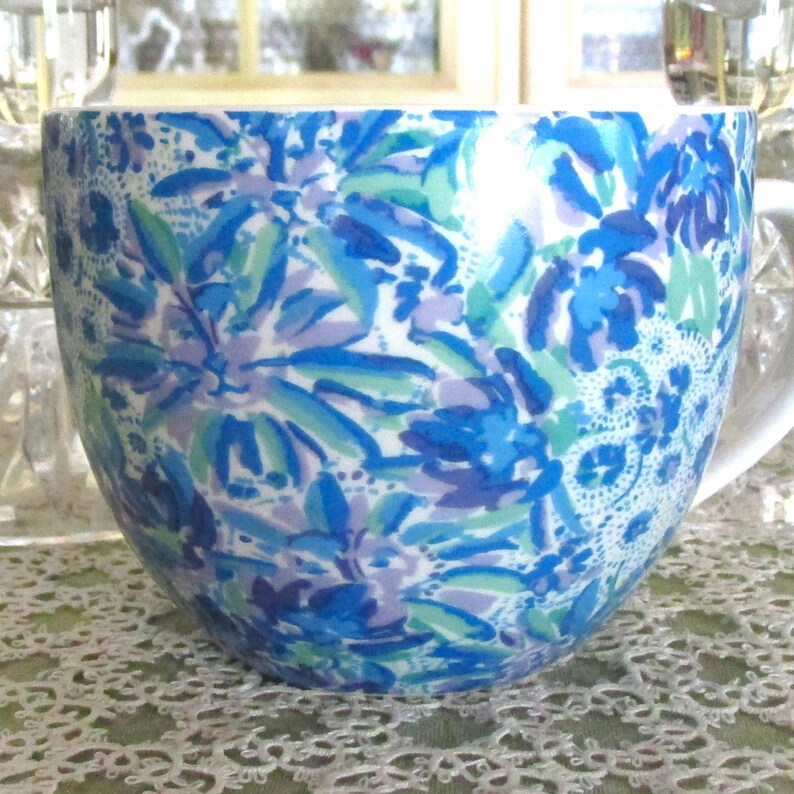 May include: A large, white ceramic mug with a vibrant floral pattern in shades of blue, green, and purple. The design features stylized flowers and leaves, creating a cheerful and artistic look. The mug has a curved shape and a white handle.