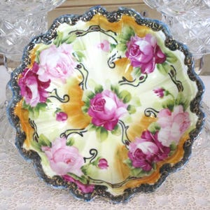 May include: A decorative, scalloped-edge dish with a floral rose design. The dish features pink and purple roses with green leaves on a yellow and orange background. The rim is trimmed with a blue and gold border.